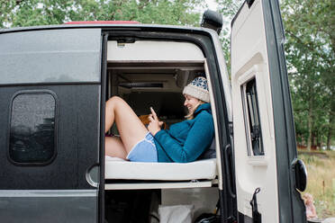 Woman sitting in bed camping in a van laughing at her cell phone - CAVF65978