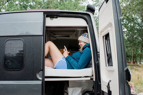 Frau im Bett sitzend, die in einem Van campiert und auf ihr Handy lacht - CAVF65978