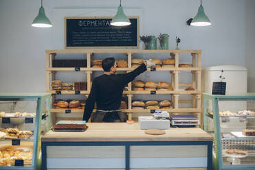 Rear view of man arranging bread on shelf in bakery - CAVF66134