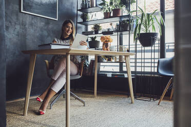 Lächelnde Geschäftsfrau, die an einem Tablet-Computer arbeitet, während sie am Tisch im Heimbüro sitzt - CAVF66151