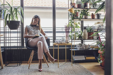 Happy businesswoman writing in diary while sitting on chair against window at home office - CAVF66158