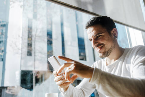 Lächelnder junger Mann arbeitet in einem Café und schaut auf sein Handy - CAVF66318