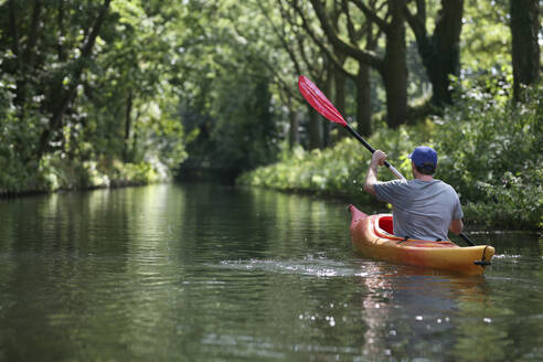 Ein kaukasischer Mann fährt mit dem Kajak einen Fluss entlang, der von einer Baumkrone beschattet wird - CAVF66386