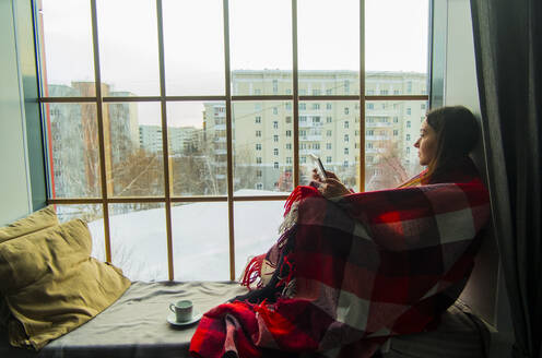 In eine Decke eingewickelte Frau, die auf einer Fensternische sitzt - CAVF66436