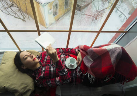 Overhead view of woman using tablet computer while relaxing on alcove window seat - CAVF66438