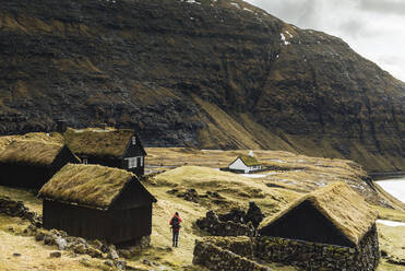 Rückansicht eines Mannes, der an Häusern auf einem Feld vor einem Berg vorbeigeht - CAVF66523