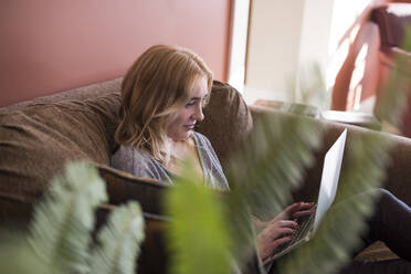 Frau benutzt einen Laptop, während sie zu Hause auf dem Sofa sitzt - CAVF66543