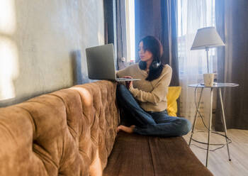 Woman using laptop computer while sitting on sofa at home - CAVF66548