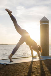 Woman practicing downward facing dog position by sea against cloudy sky during sunset - CAVF66612