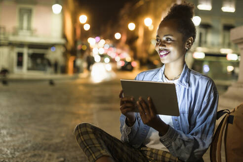 Porträt einer lächelnden jungen Frau mit digitalem Tablet in der Stadt bei Nacht, Lissabon, Portugal - UUF19128