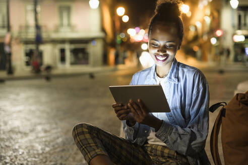 Porträt einer glücklichen jungen Frau, die ein digitales Tablet in der nächtlichen Stadt benutzt, Lissabon, Portugal - UUF19129
