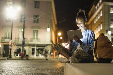 Porträt einer lächelnden jungen Frau, die ein digitales Tablet in der nächtlichen Stadt benutzt, Lissabon, Portugal - UUF19131