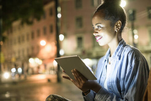 Porträt einer lächelnden jungen Frau mit digitalem Tablet in der Stadt bei Nacht, Lissabon, Portugal - UUF19133