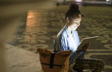 Glückliche junge Frau mit digitalem Tablet auf einem Platz in der nächtlichen Stadt, Lissabon, Portugal - UUF19134