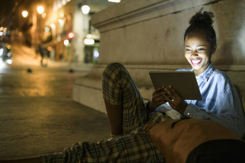 Porträt einer glücklichen jungen Frau, die ein digitales Tablet in der nächtlichen Stadt benutzt, Lissabon, Portugal, lizenzfreies Stockfoto