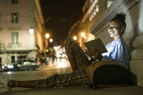 Porträt einer glücklichen jungen Frau mit Rucksack und digitalem Tablet in der nächtlichen Stadt, Lissabon, Portugal - UUF19136