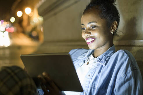 Porträt einer glücklichen jungen Frau, die ein digitales Tablet in der nächtlichen Stadt benutzt, Lissabon, Portugal - UUF19137