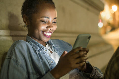 Porträt einer glücklichen jungen Frau, die ihr Smartphone in der nächtlichen Stadt benutzt, Lissabon, Portugal, lizenzfreies Stockfoto