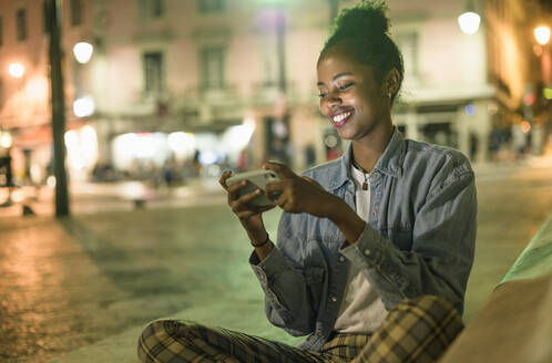 Porträt einer glücklichen jungen Frau, die ihr Smartphone in der nächtlichen Stadt benutzt, Lissabon, Portugal - UUF19144