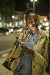 Porträt einer entspannten jungen Frau mit Kopfhörern und Smartphone in der nächtlichen Stadt, Lissabon, Portugal - UUF19147
