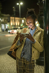 Portrait of smiling young woman using earphones and smartphone in the city by night, Lisbon, Portugal - UUF19148