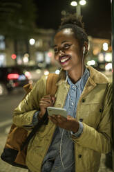 Portrait of happy young woman using earphones and smartphone in the city by night, Lisbon, Portugal - UUF19149