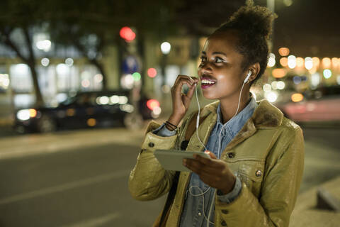 Portrait of smiling young woman using earphones and smartphone in the city by night, Lisbon, Portugal stock photo