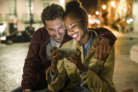 Porträt eines glücklichen jungen Paares, das gemeinsam ein Smartphone bei Nacht betrachtet, Lissabon, Portugal, lizenzfreies Stockfoto