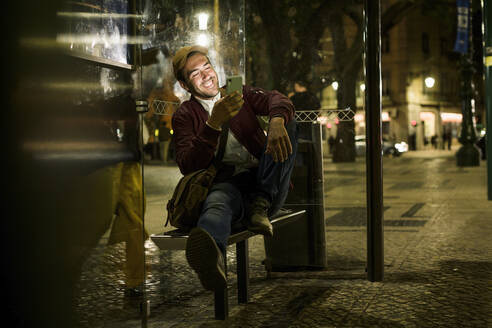 Porträt eines lachenden jungen Mannes, der nachts an einer Bushaltestelle sitzt und sein Smartphone benutzt, Lissabon, Portugal - UUF19175