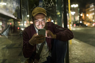 Porträt eines lächelnden jungen Mannes, der an einer nächtlichen Bushaltestelle sitzt und ein Mobiltelefon benutzt, Lissabon, Portugal - UUF19179