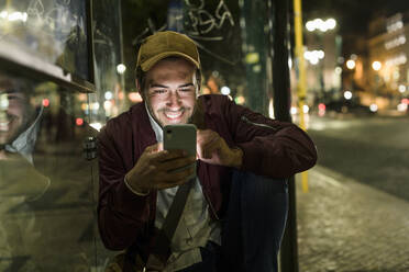 Portrait of smiling young man sitting at bus stop by night using cell phone, Lisbon, Portugal - UUF19179