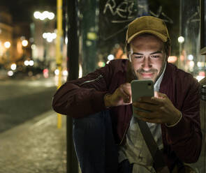 Portrait of smiling young man sitting at bus stop by night using cell phone, Lisbon, Portugal - UUF19180