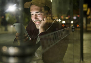 Portrait of smiling young man sitting at bus stop by night looking at cell phone, Lisbon, Portugal - UUF19181