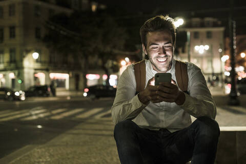Porträt eines glücklichen jungen Mannes, der ein Handy in der nächtlichen Stadt benutzt, Lissabon, Portugal, lizenzfreies Stockfoto