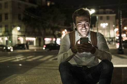 Porträt eines glücklichen jungen Mannes, der ein Handy in der nächtlichen Stadt benutzt, Lissabon, Portugal - UUF19186
