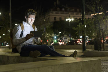 Portrait of young man using digital tablet and earphones in the city by night, Lisbon, Portugal - UUF19189