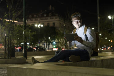 Portrait of young man using digital tablet and earphones in the city by night, Lisbon, Portugal - UUF19191