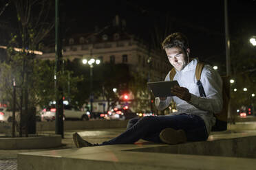 Portrait of young man using digital tablet and earphones in the city by night, Lisbon, Portugal - UUF19192