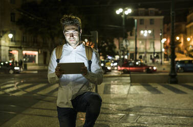 Portrait of young man using digital tablet and earphones in the city by night, Lisbon, Portugal - UUF19193