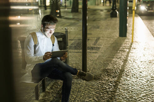 Porträt eines lachenden jungen Mannes, der an einer Bushaltestelle sitzt und ein digitales Tablet und Kopfhörer benutzt, Lissabon, Portugal, bei Nacht - UUF19196