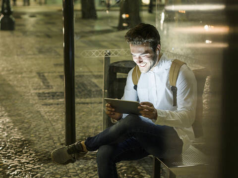 Porträt eines glücklichen jungen Mannes, der an einer Bushaltestelle sitzt und ein digitales Tablet und Kopfhörer benutzt, Lissabon, Portugal, bei Nacht, lizenzfreies Stockfoto