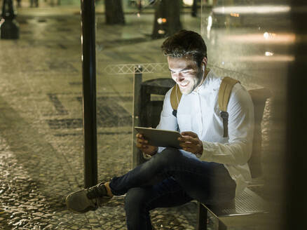 Porträt eines glücklichen jungen Mannes, der an einer Bushaltestelle sitzt und ein digitales Tablet und Kopfhörer benutzt, Lissabon, Portugal, bei Nacht - UUF19197