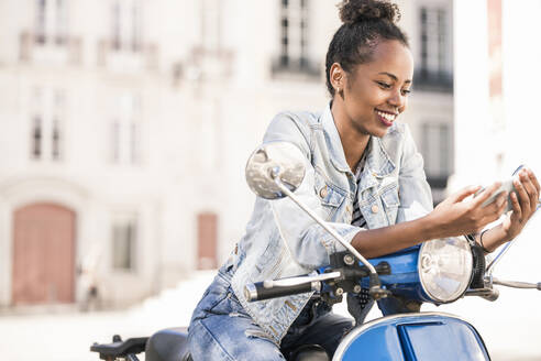 Glückliche junge Frau mit Motorroller und Mobiltelefon in der Stadt, Lissabon, Portugal - UUF19202