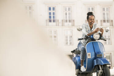 Smiling young woman with motor scooter and mobile phone in the city, Lisbon, Portugal - UUF19205