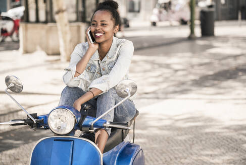 Glückliche junge Frau mit Motorroller beim Telefonieren in der Stadt, Lissabon, Portugal - UUF19206