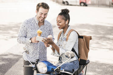 Happy young couple with motor scooter and ice cream using mobile phone in the city, Lisbon, Portugal - UUF19208
