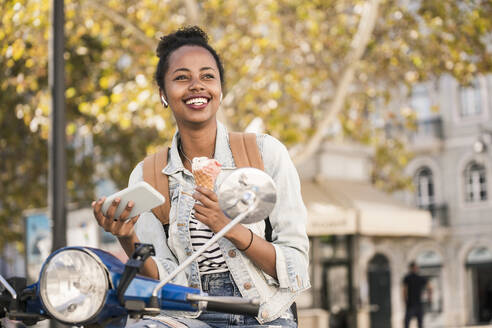 Glückliche junge Frau mit Motorroller beim Eisessen in der Stadt, Lissabon, Portugal - UUF19213