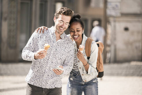 Glückliches junges Paar mit Eis und Mobiltelefon in der Stadt unterwegs, Lissabon, Portugal - UUF19215