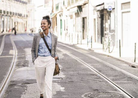 Smiling young woman with wireless earphones in the city on the go, Lisbon, Portugal - UUF19228