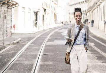 Smiling young woman with mobile phone in the city on the go, Lisbon, Portugal - UUF19229